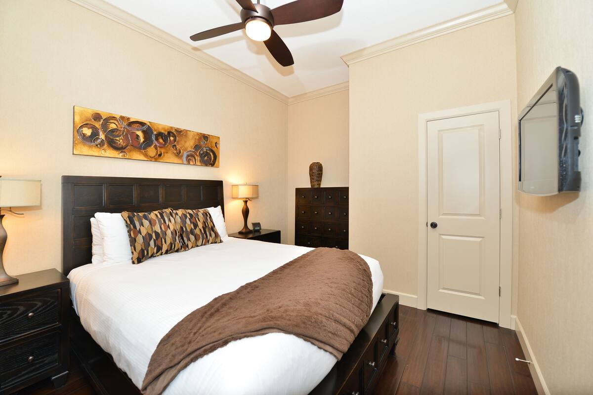 A hotel room with a bed, a ceiling fan, a painting, a dresser, and a door.