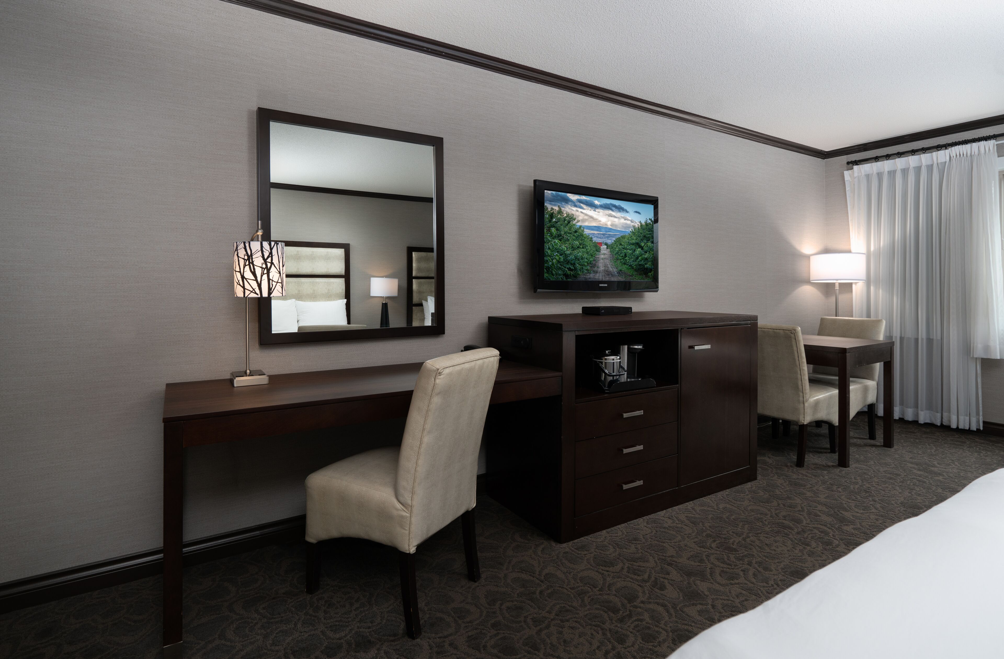 A hotel room with a large bed, a desk with a chair, a television, a dresser, and a lamp.
