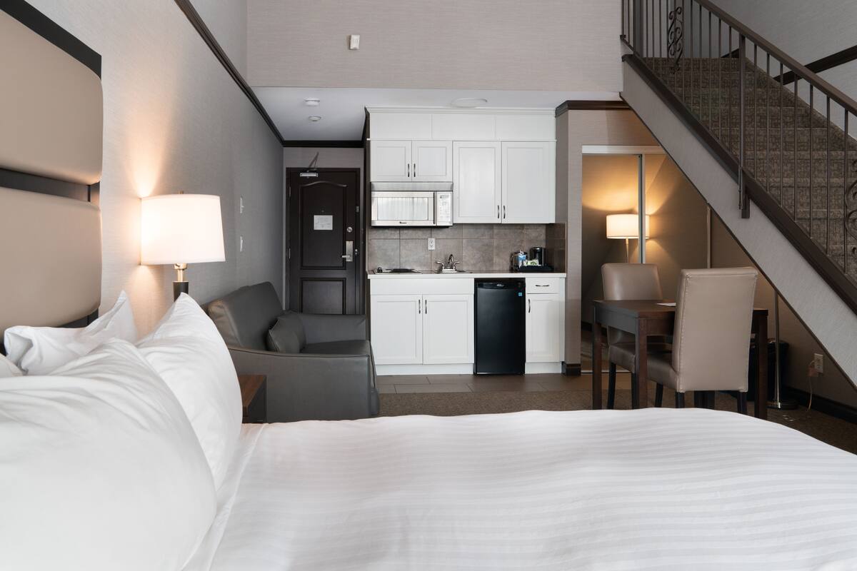 A hotel room with a bed, a desk, a chair, a kitchenette, and a staircase.