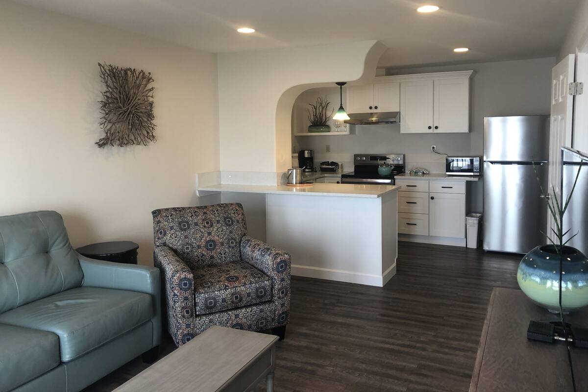A modern kitchen with white cabinets, a stainless steel refrigerator, and a microwave. There is a patterned armchair and a grey sofa in the living area.