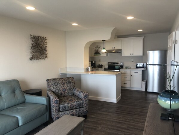 A modern kitchen with white cabinets, a stainless steel refrigerator, and a microwave. There is a patterned armchair and a grey sofa in the living area.