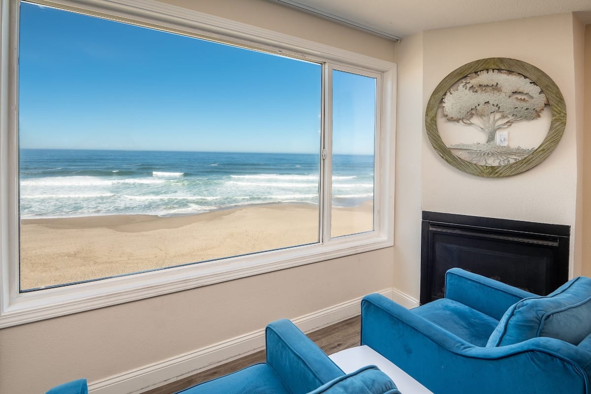 A room with a large window overlooking a beach, a fireplace, and blue upholstered furniture.