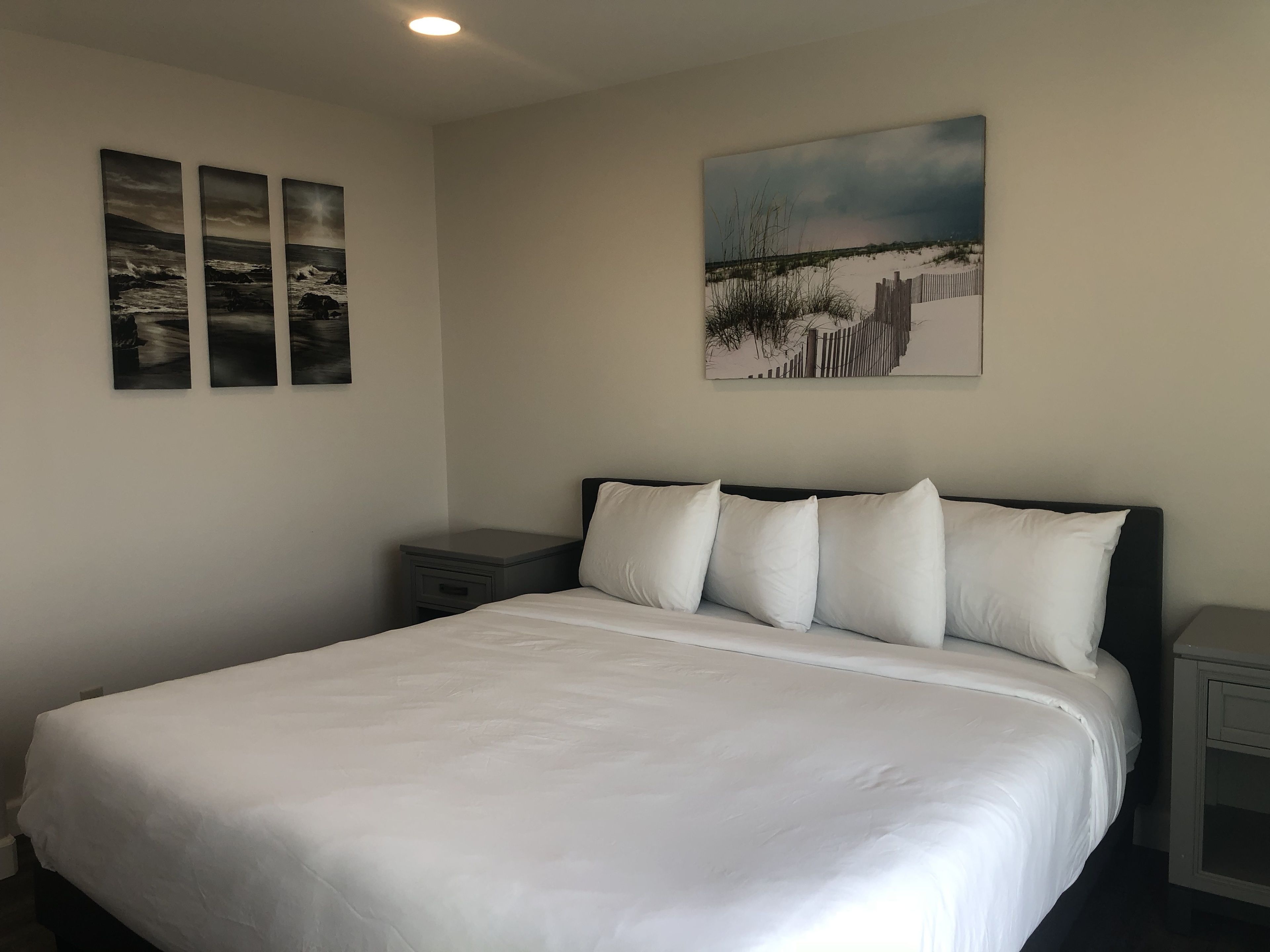 A neatly made bed with white linens, a dark headboard, and a nightstand with a drawer. There are three framed pictures on the wall above the bed.