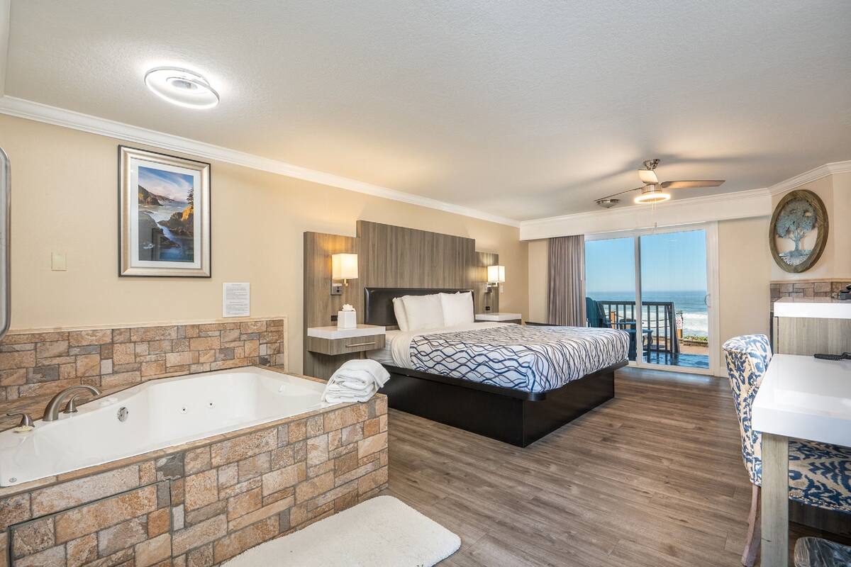 A hotel room with a large bed, a bathtub, a balcony with a view of the ocean, and a dining area.