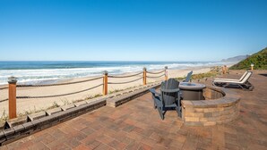 A beachside patio with a view of the ocean, a seating area with chairs and a fire pit, and a rope barrier along the shoreline.
