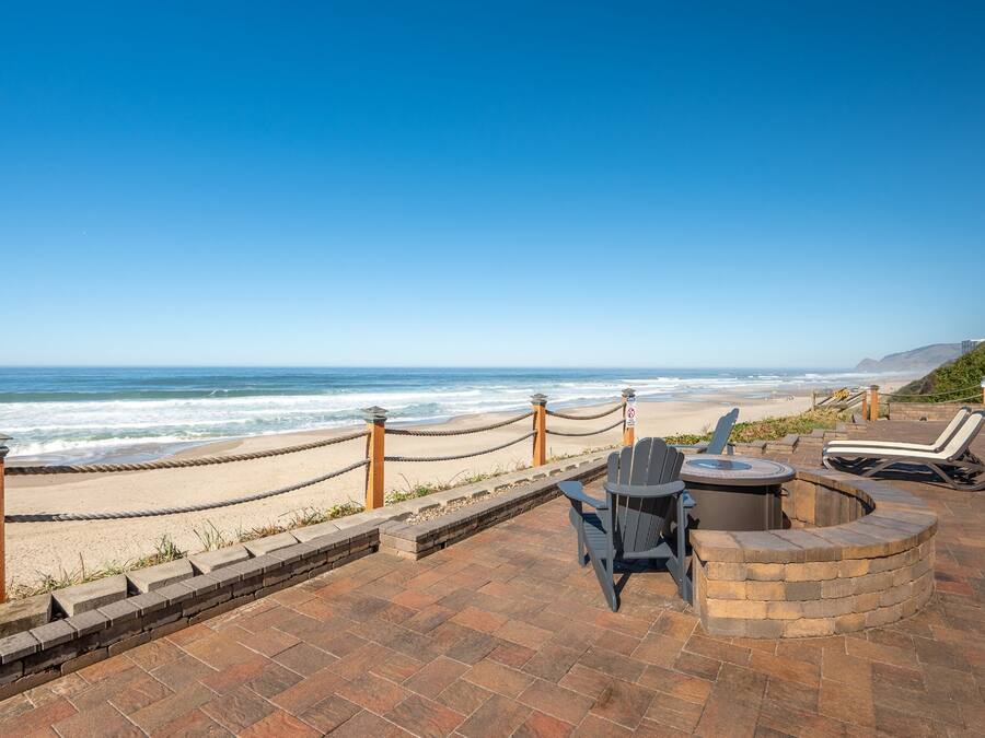 A beachside patio with a view of the ocean, a seating area with chairs and a fire pit, and a rope barrier along the shoreline.
