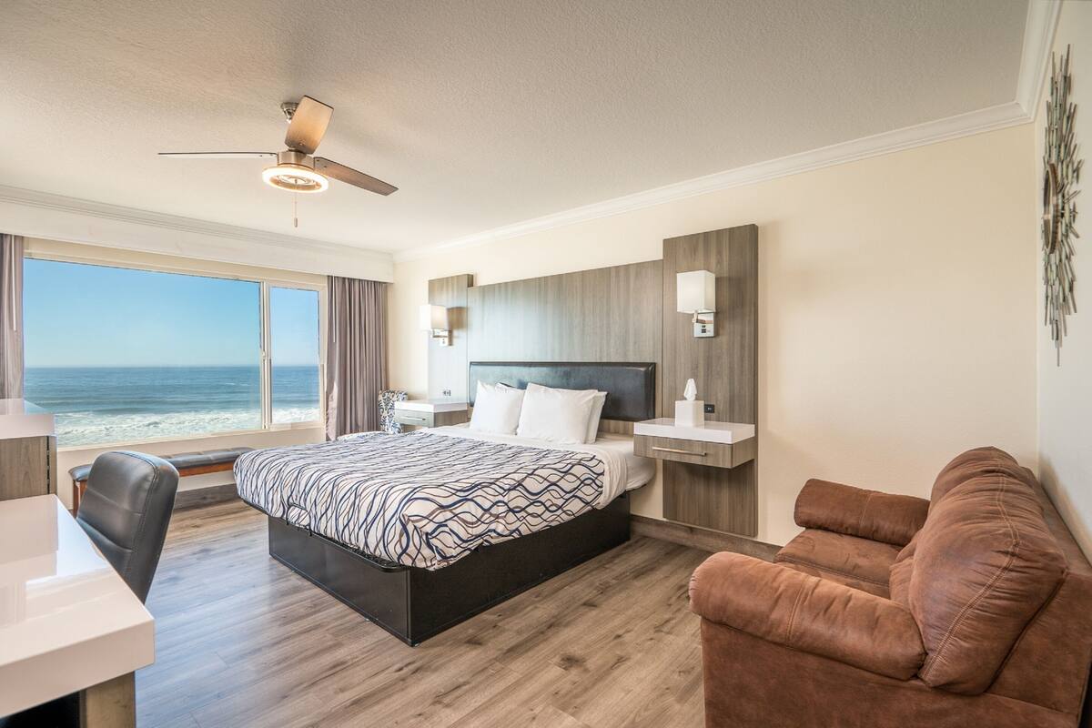 A modern hotel room with a large bed, a desk, a brown leather sofa, and a view of the ocean.