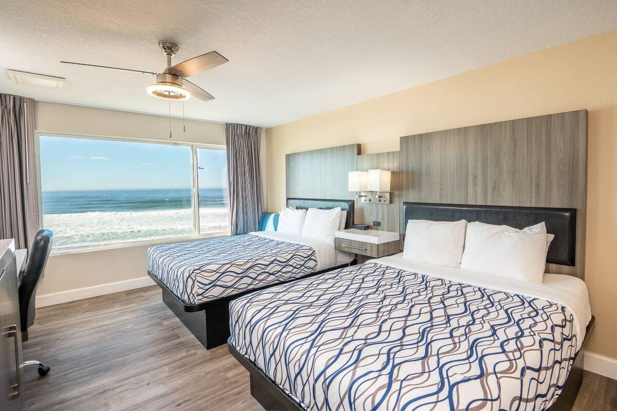 A hotel room with two beds, a ceiling fan, a large window with a view of the ocean, and a desk with a chair.