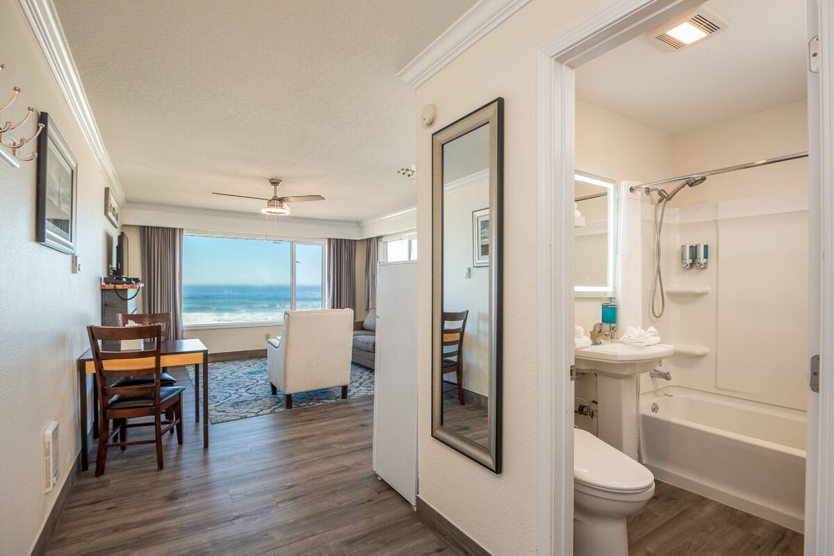 A hotel room with a view of the ocean, a dining area with a table and chairs, a large mirror, and a bathroom with a bathtub and toilet.