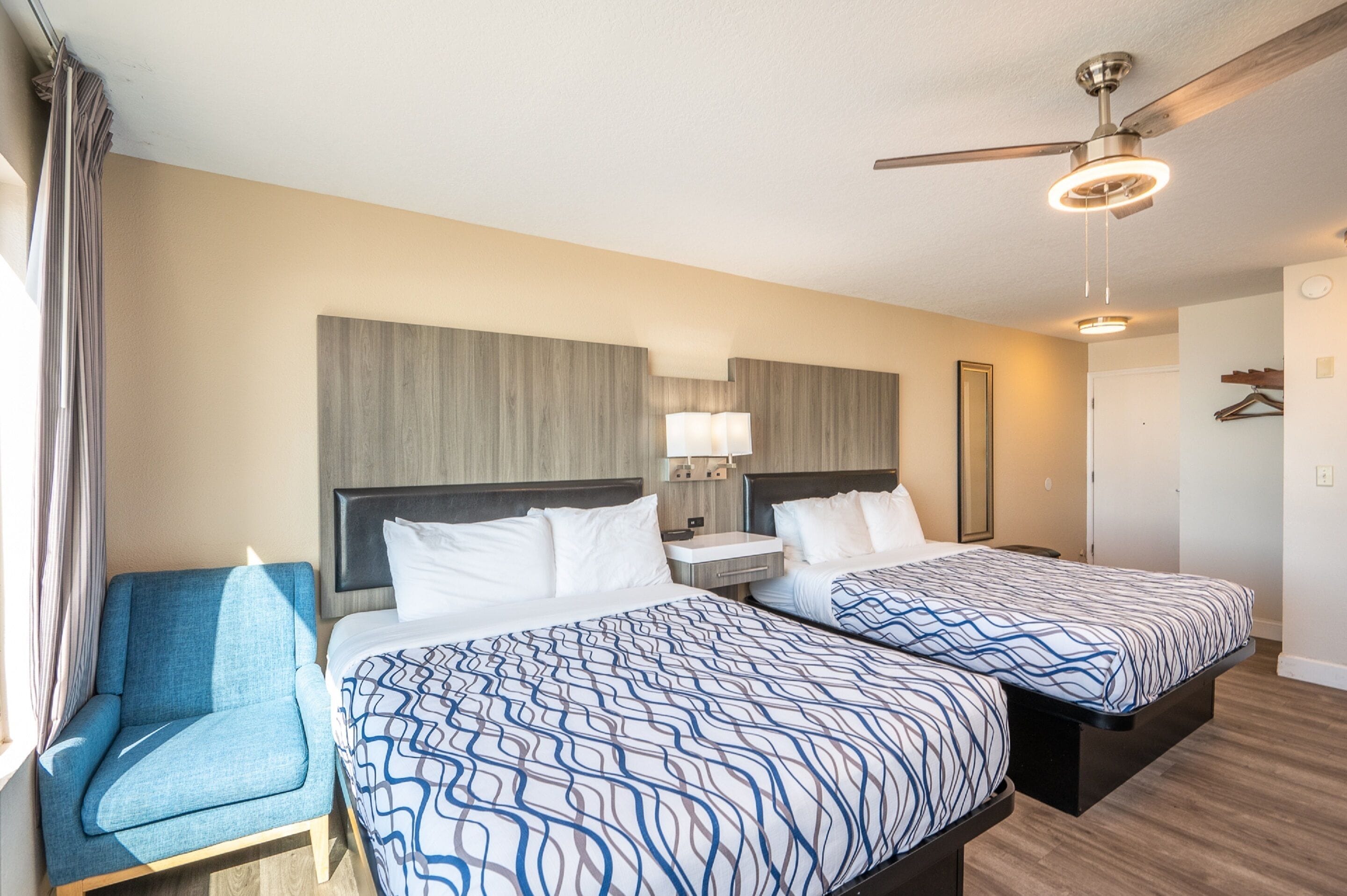Une chambre d’hôtel avec deux lits, une chaise, un ventilateur de plafond et une table de chevet avec une lampe.
