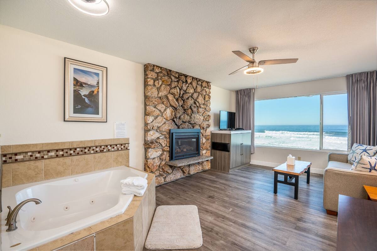 A modern bathroom with a bathtub, a stone accent wall, a mounted TV, and a view of the ocean through large windows.