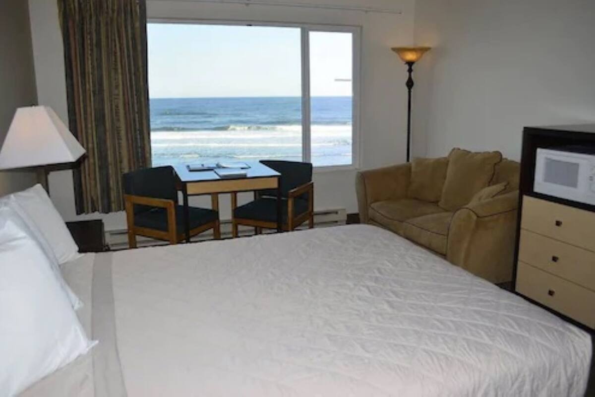 A hotel room with a bed, a desk with two chairs, a sofa, a lamp, and a view of the ocean.
