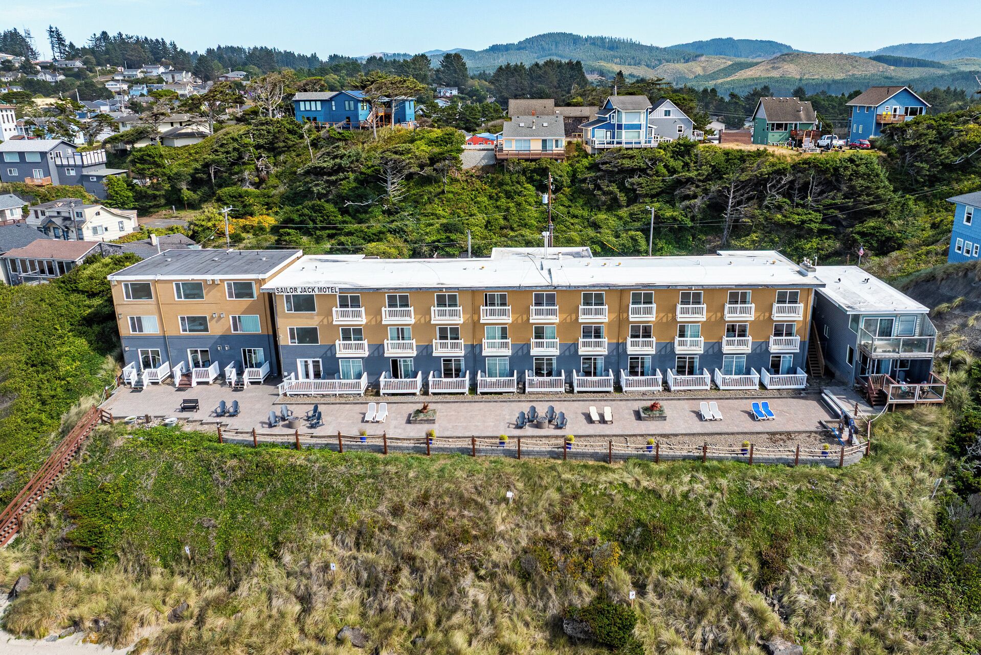 A multi-story motel with a flat roof, a parking area with sun loungers, and a view of the surrounding residential area and hills.