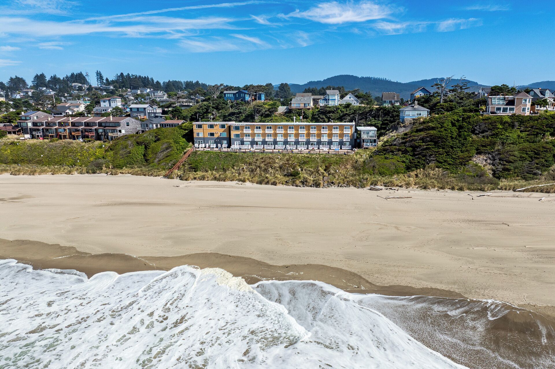 A coastal hotel with multiple buildings, a sandy beach, and a forested hillside in the background.