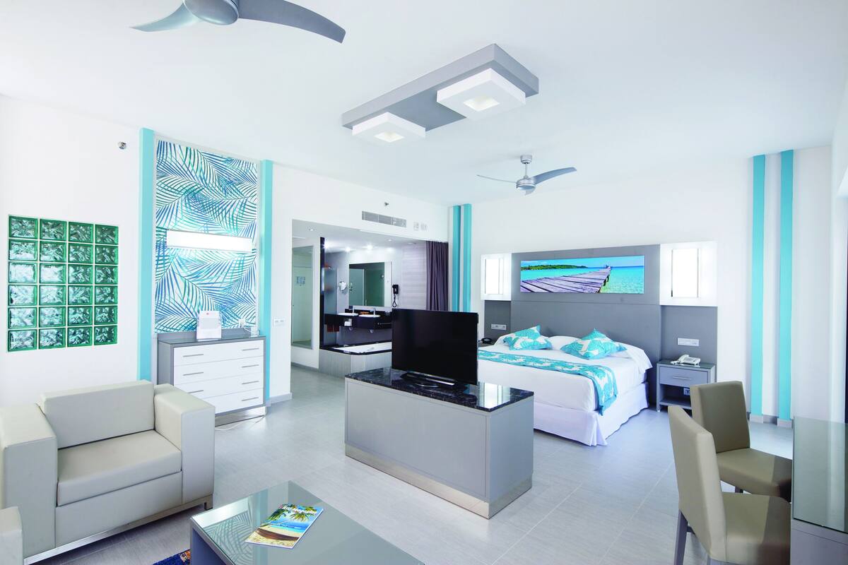 A modern hotel room with a flat-screen TV, a sofa, a coffee table, a bed with a blue and white bedspread, a desk, and a chair.