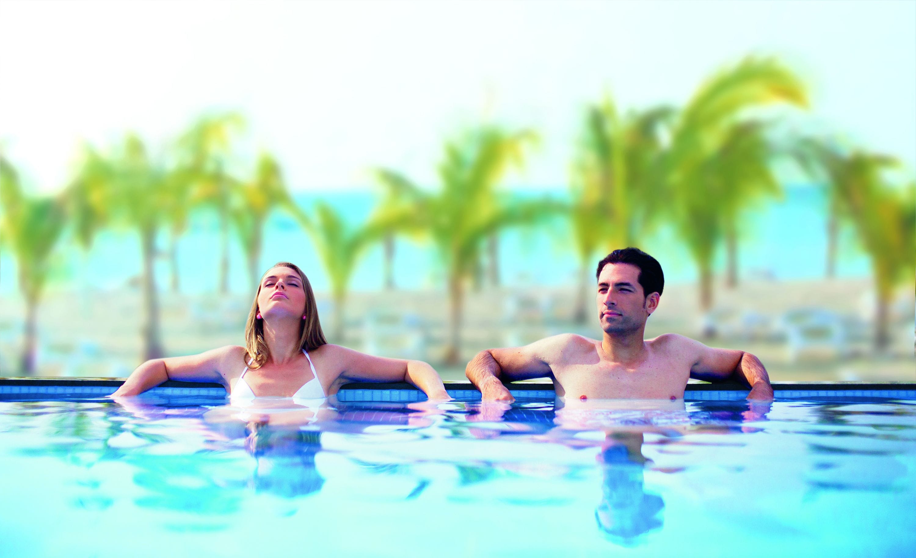 Two people in a swimming pool with a view of palm trees and a beach in the background.