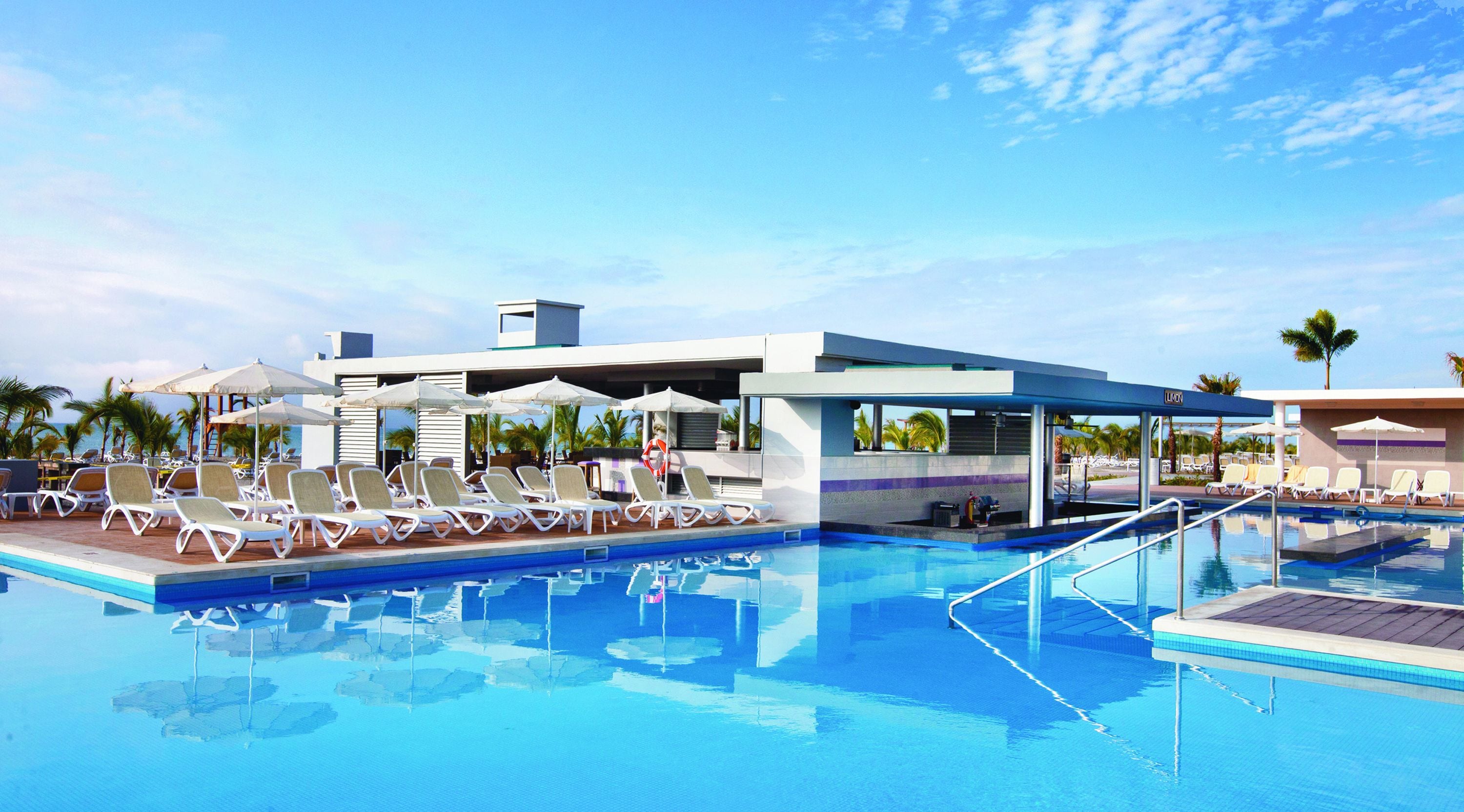 Ein Swimmingpool mit Liegestühlen und Sonnenschirmen, umgeben von Palmen und blauem Himmel.