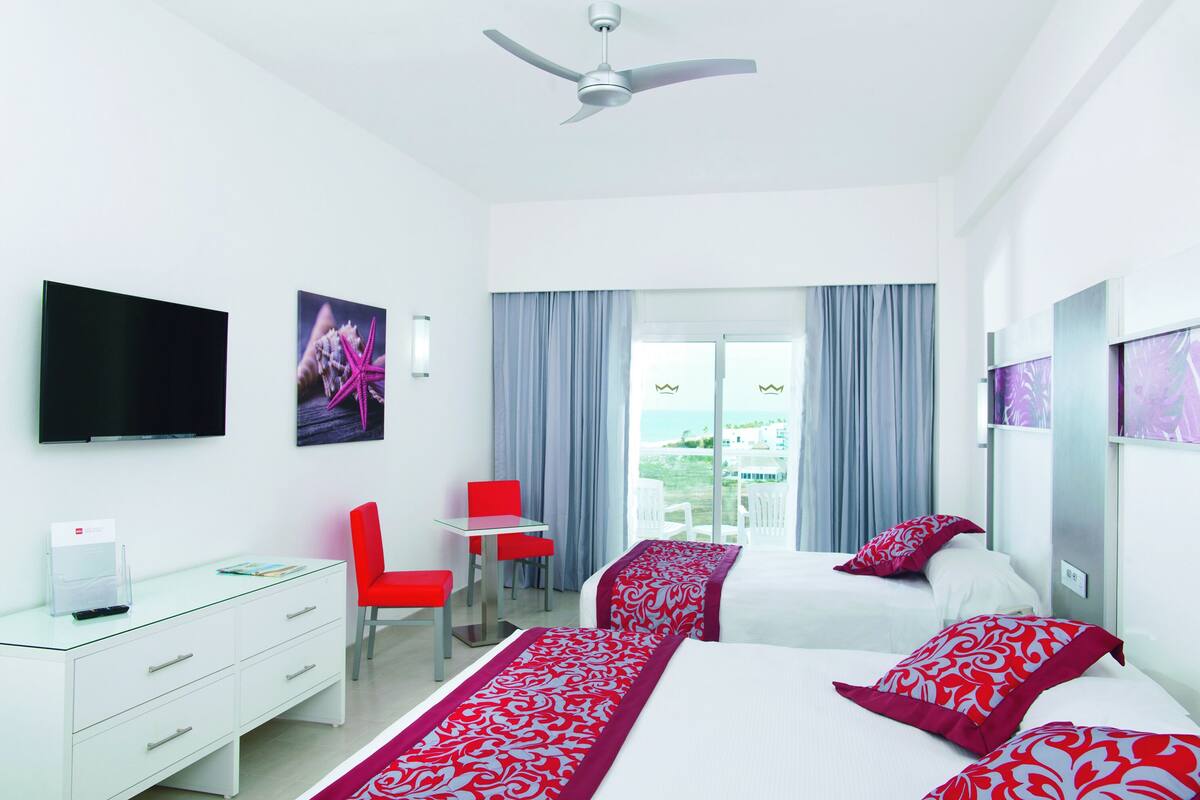 A hotel room with two beds, a desk, a chair, a TV, and a balcony with curtains.