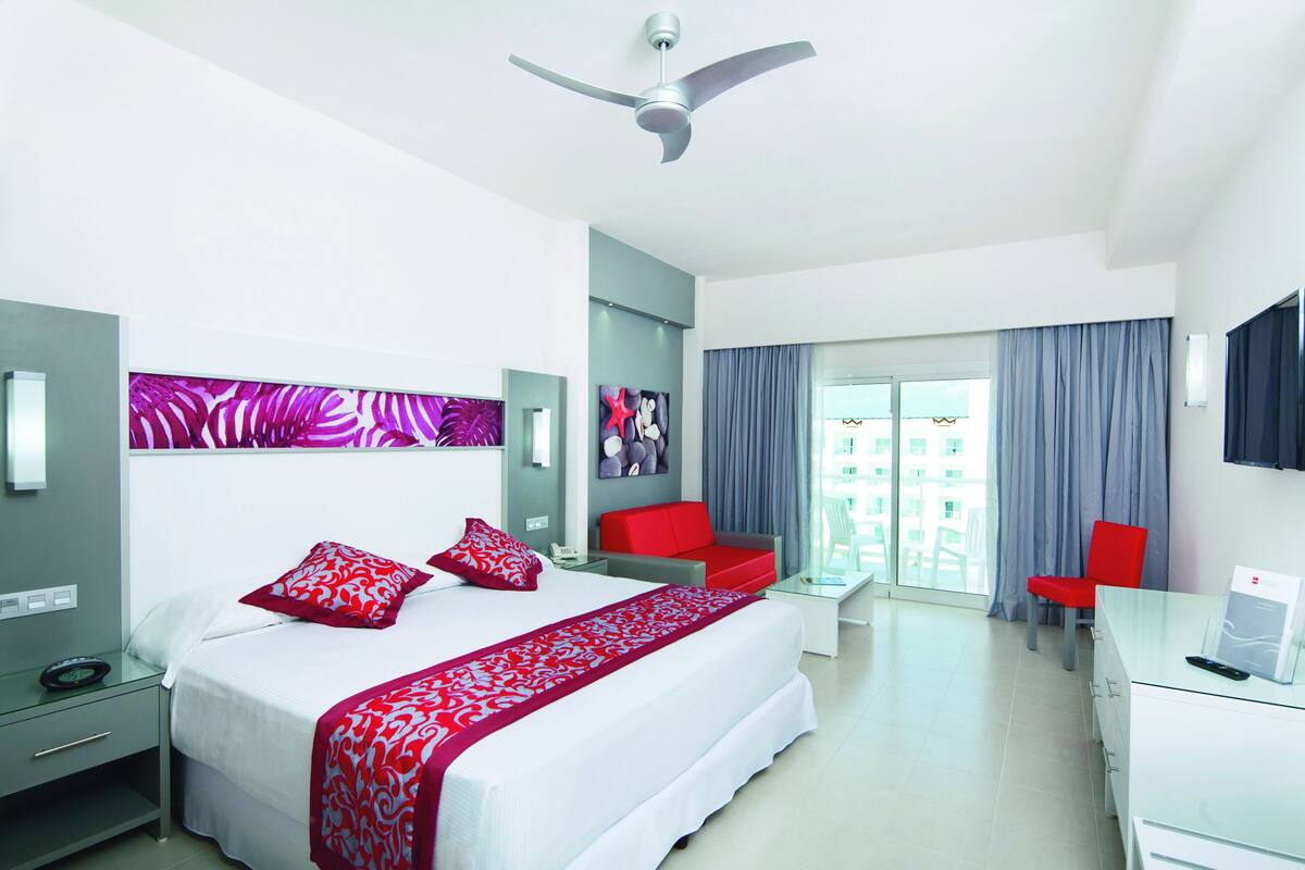 A modern hotel room with a large bed, a desk, a chair, and a ceiling fan.