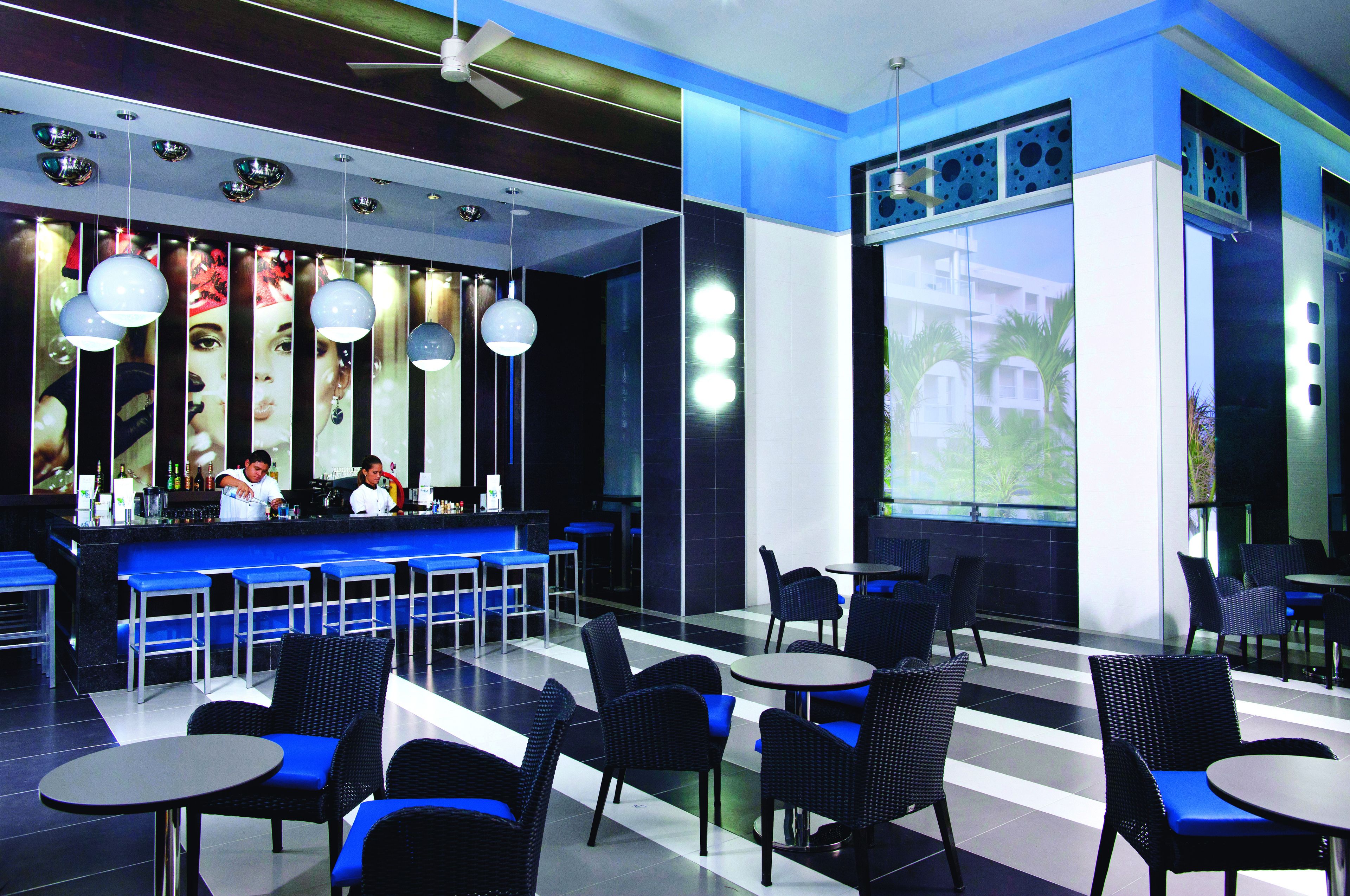 A modern restaurant with blue bar stools, black wicker chairs, and a large window offering a view of palm trees.