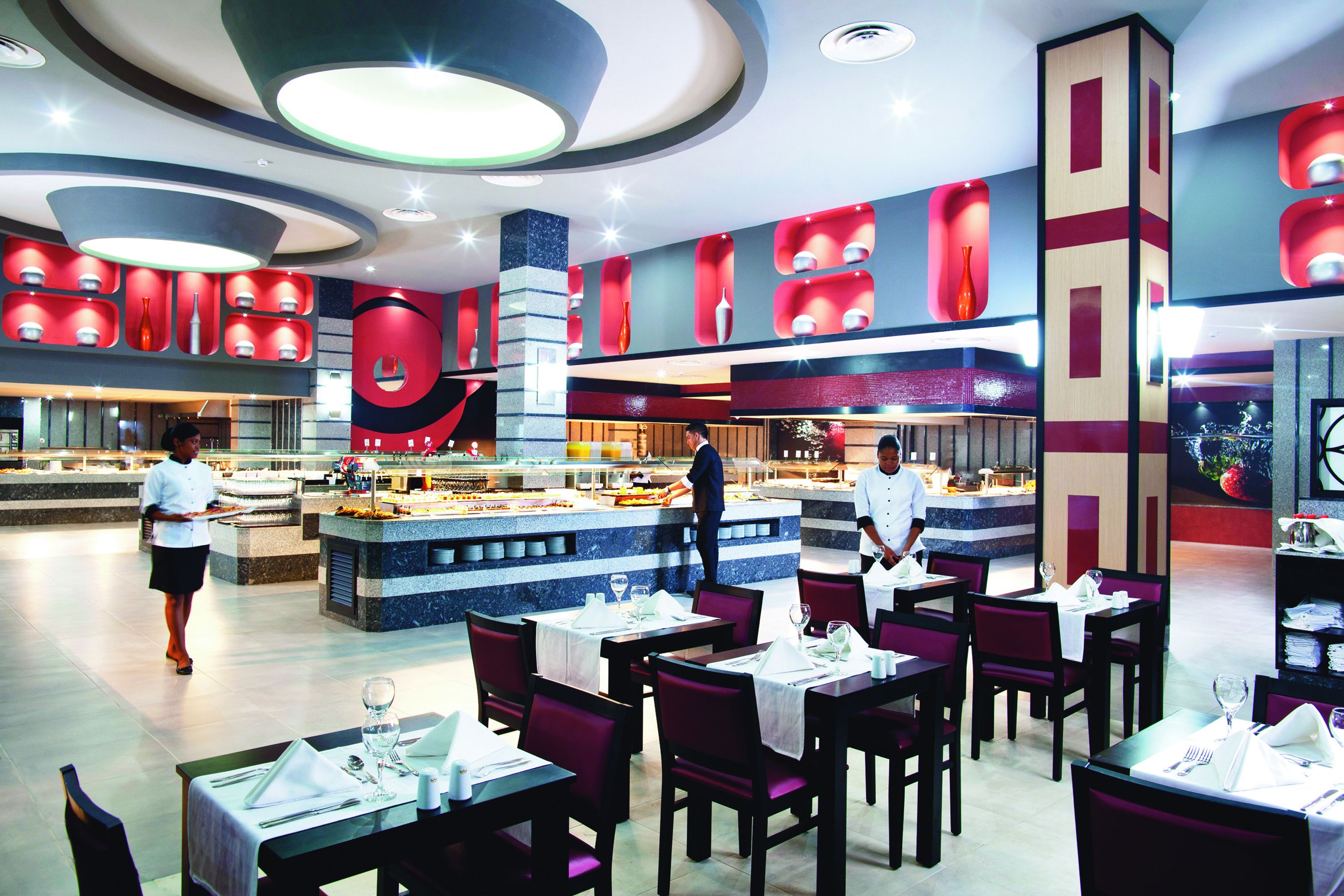 A modern restaurant with a central bar area, multiple dining tables, and a waiter serving.