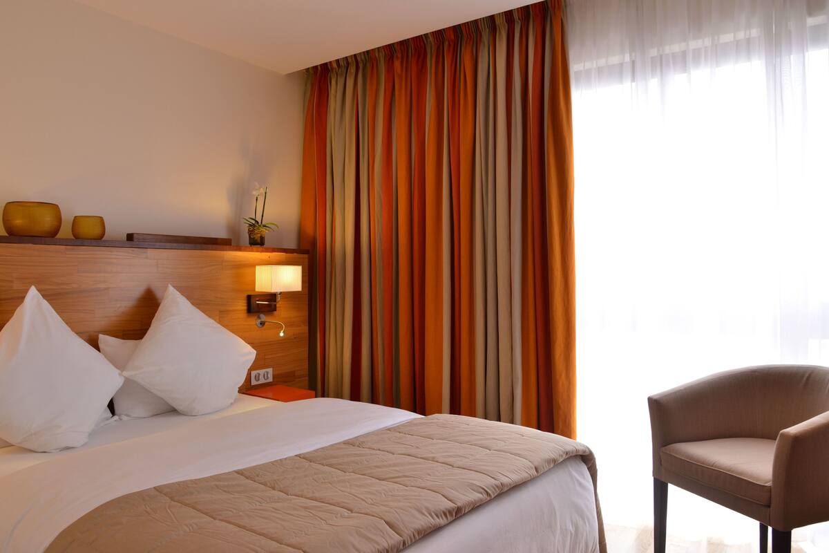 A hotel room with a bed, a nightstand, a lamp, a chair, and a window with curtains.
