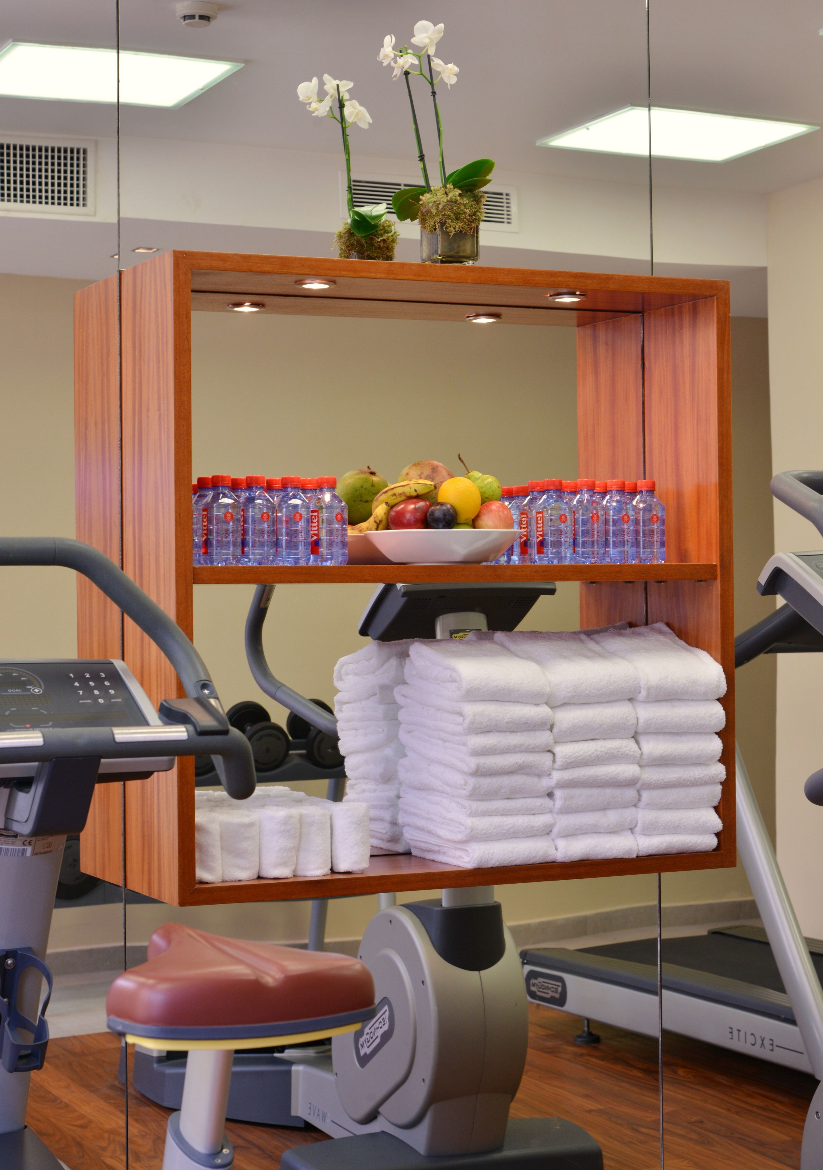 A fitness room with exercise equipment, a shelf with towels, a bowl of fruit, and bottled water.