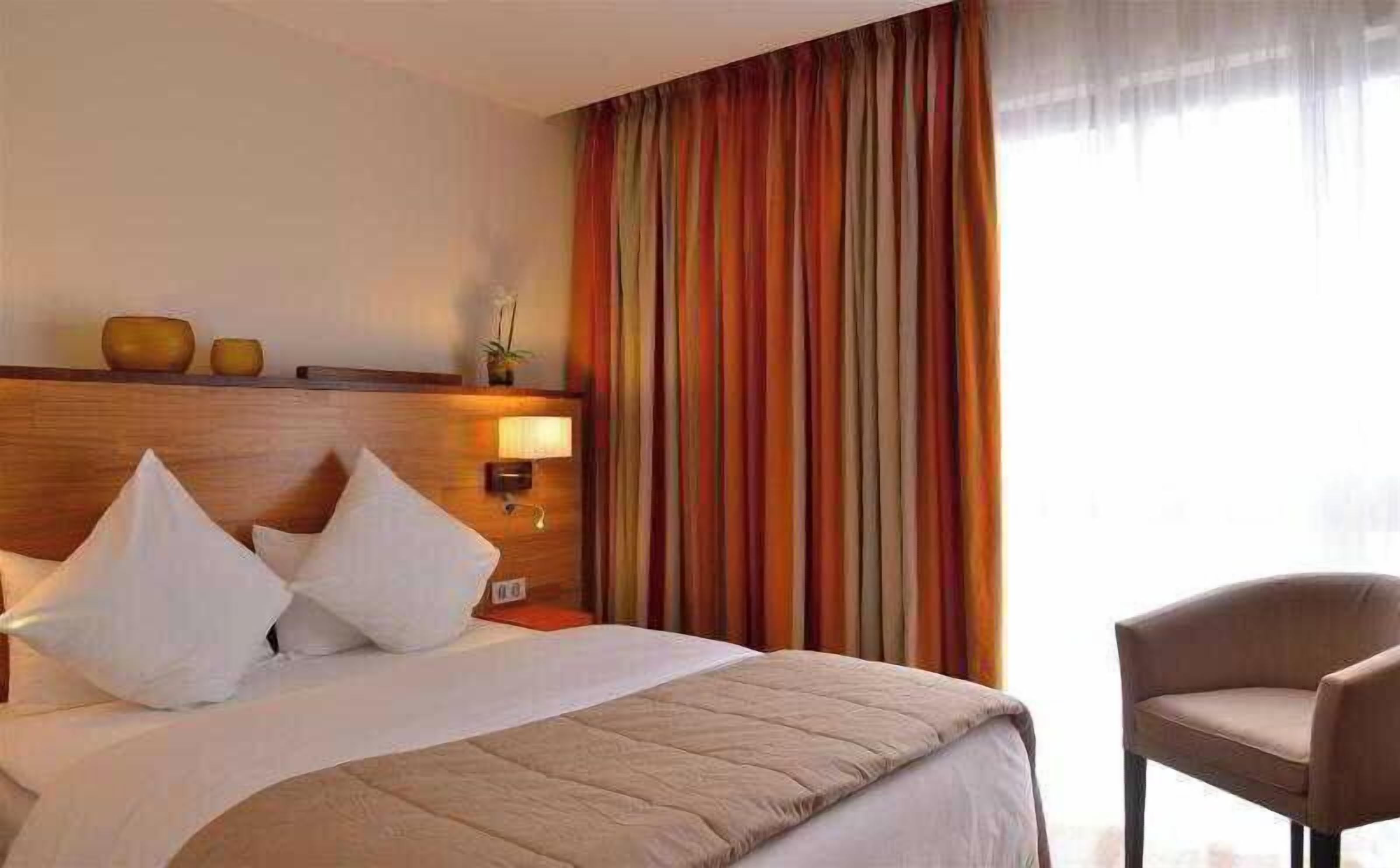 Quarto de hotel com cama, almofadas, mesa de cabeceira, luminária, cadeira e janela com cortinas.