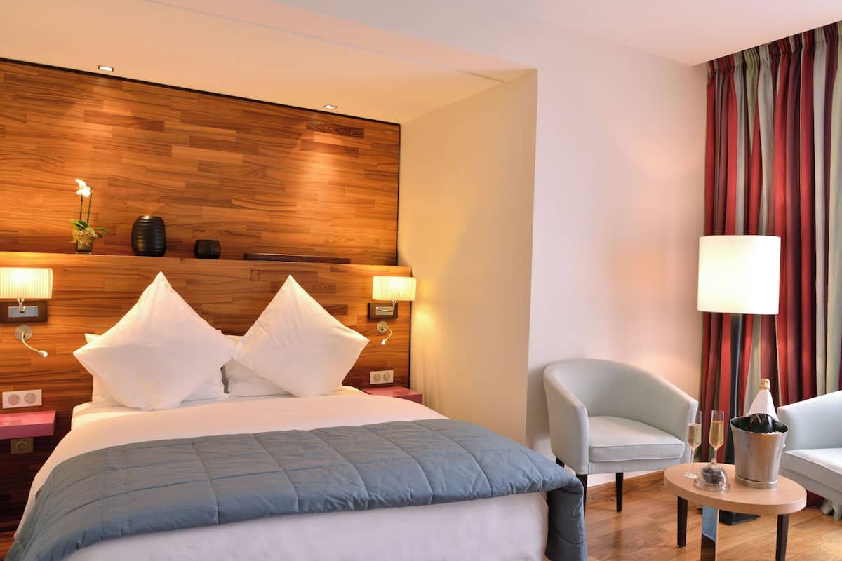 A hotel room with a wooden headboard, two bedside lamps, a bed with white and blue bedding, a light-colored armchair, a small round table with a lamp, and a window with red curtains.