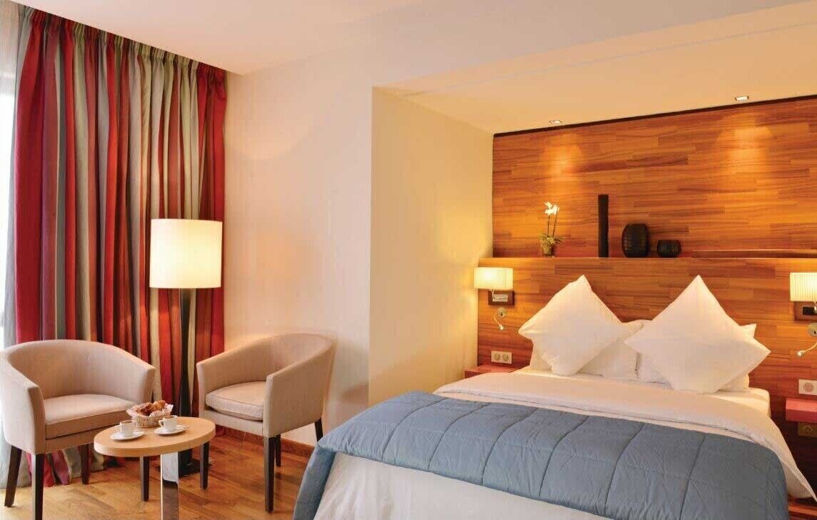 Quarto de hotel com cama, duas cadeiras, mesa pequena com abajur e cabeceira de madeira.