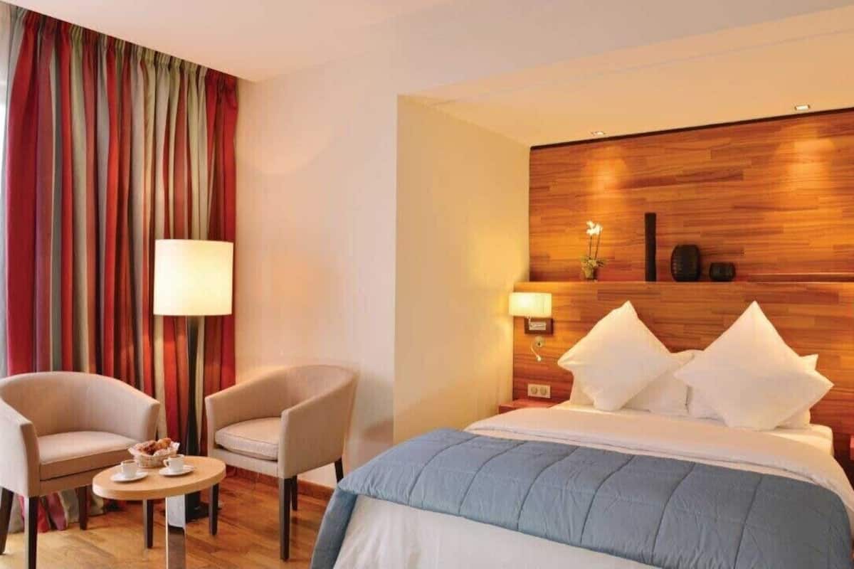 A hotel room with a bed, two chairs, a small table with a lamp, and a wooden headboard.