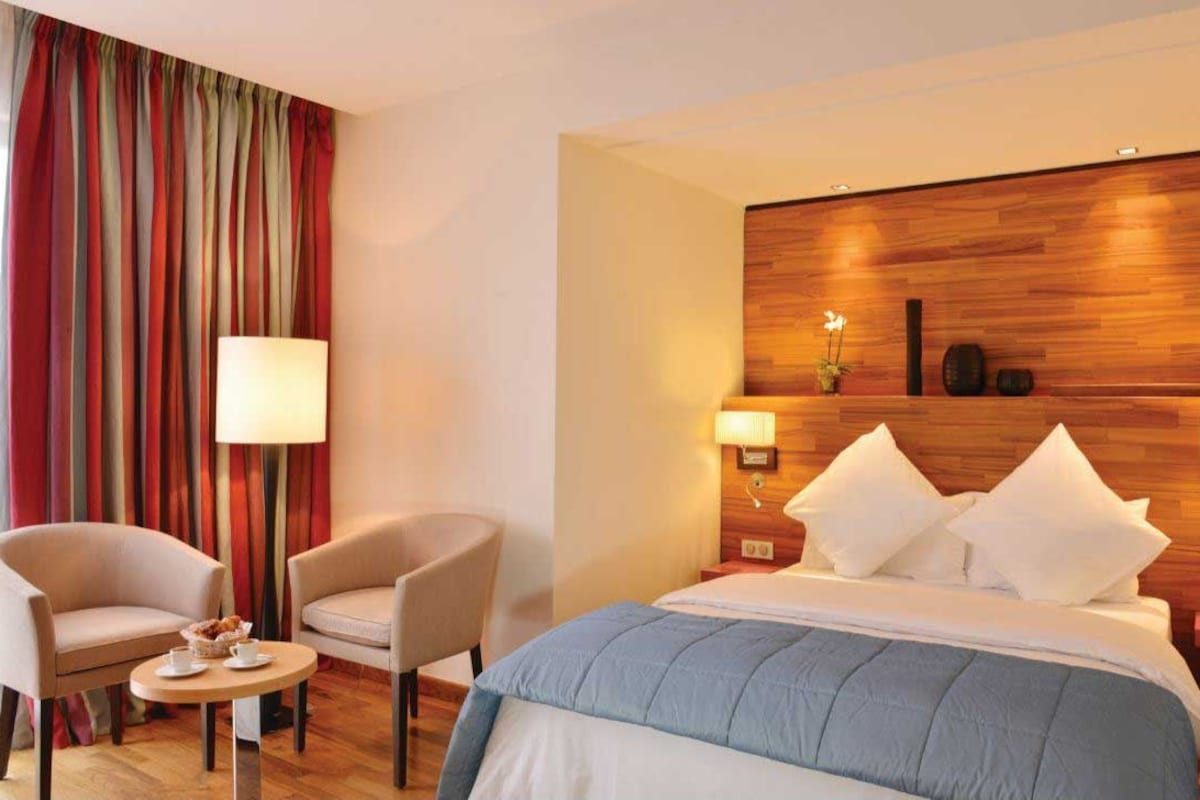 A hotel room with a bed, two chairs, a small table with a lamp, and a wooden headboard.
