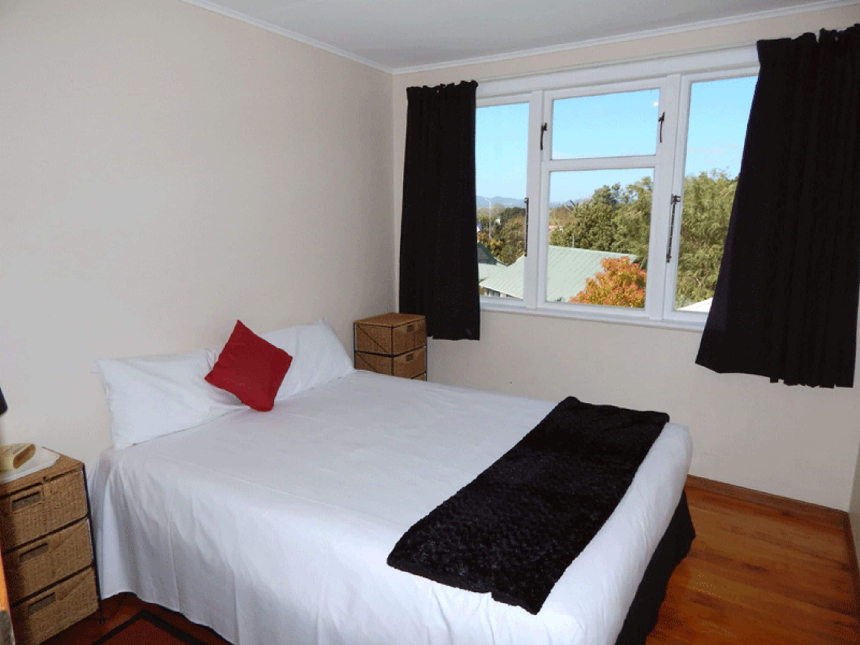 Un dormitorio con cama, ventana con cortinas, mesita de noche y una caja.