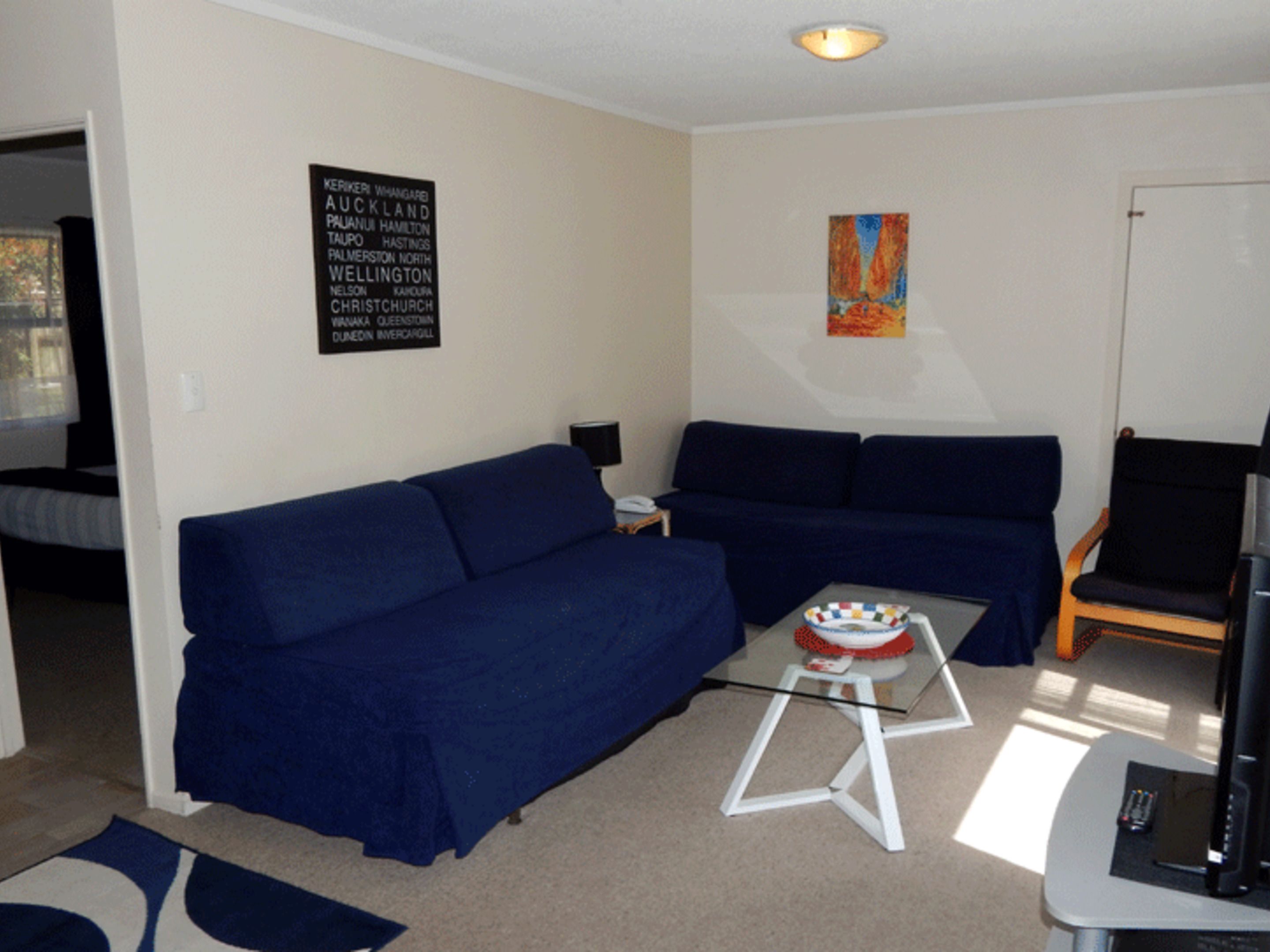 A living room with a blue sofa, a glass coffee table, a television, and a framed sign listing New Zealand cities.