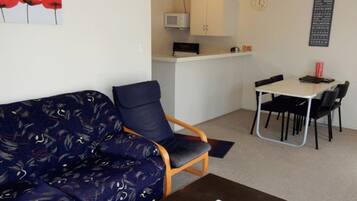A living room with a sofa, a dining table with chairs, a kitchen area with a microwave, and a clock on the wall.