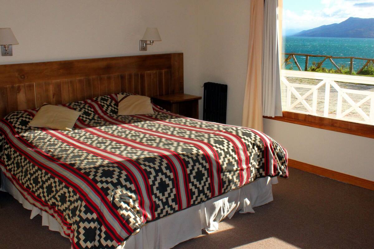 Un dormitorio con una cama grande, dos mesitas de noche y vistas a un lago y montañas a través de una ventana.
