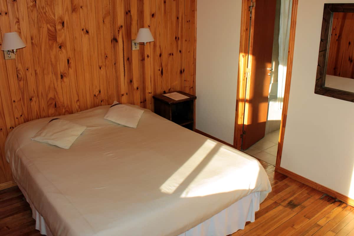 Un dormitorio con pared de madera, una cama con colcha blanca, dos lámparas de noche y una mesita auxiliar pequeña.