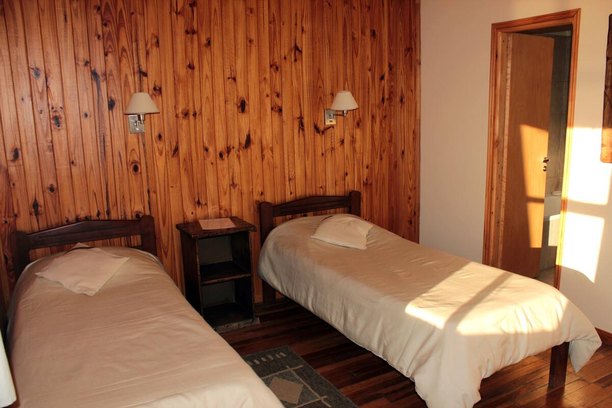 Una habitación con dos camas de madera, una mesita de noche y una lámpara fijada a la pared.