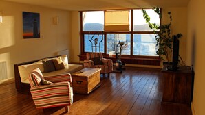 Amplia sala de estar con piso de madera, un sofá, sillones a rayas, una mesita y un televisor. Grandes ventanales ofrecen vista al mar.