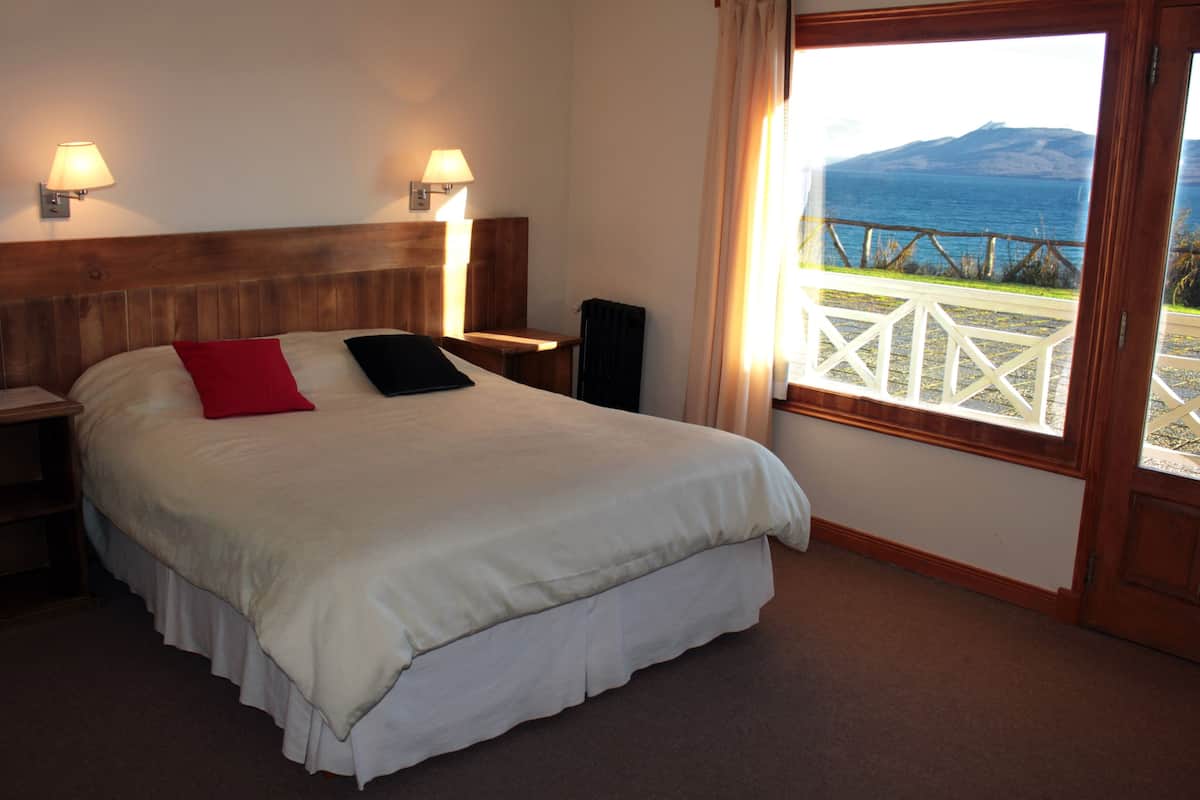 Una cama con una colcha blanca, dos almohadas, una mesita de noche con una lámpara, un teléfono y un altavoz. Se ve una ventana con vistas al mar y a las montañas.