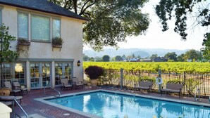 A pool area with a brick patio, outdoor seating, and a view of a vineyard.