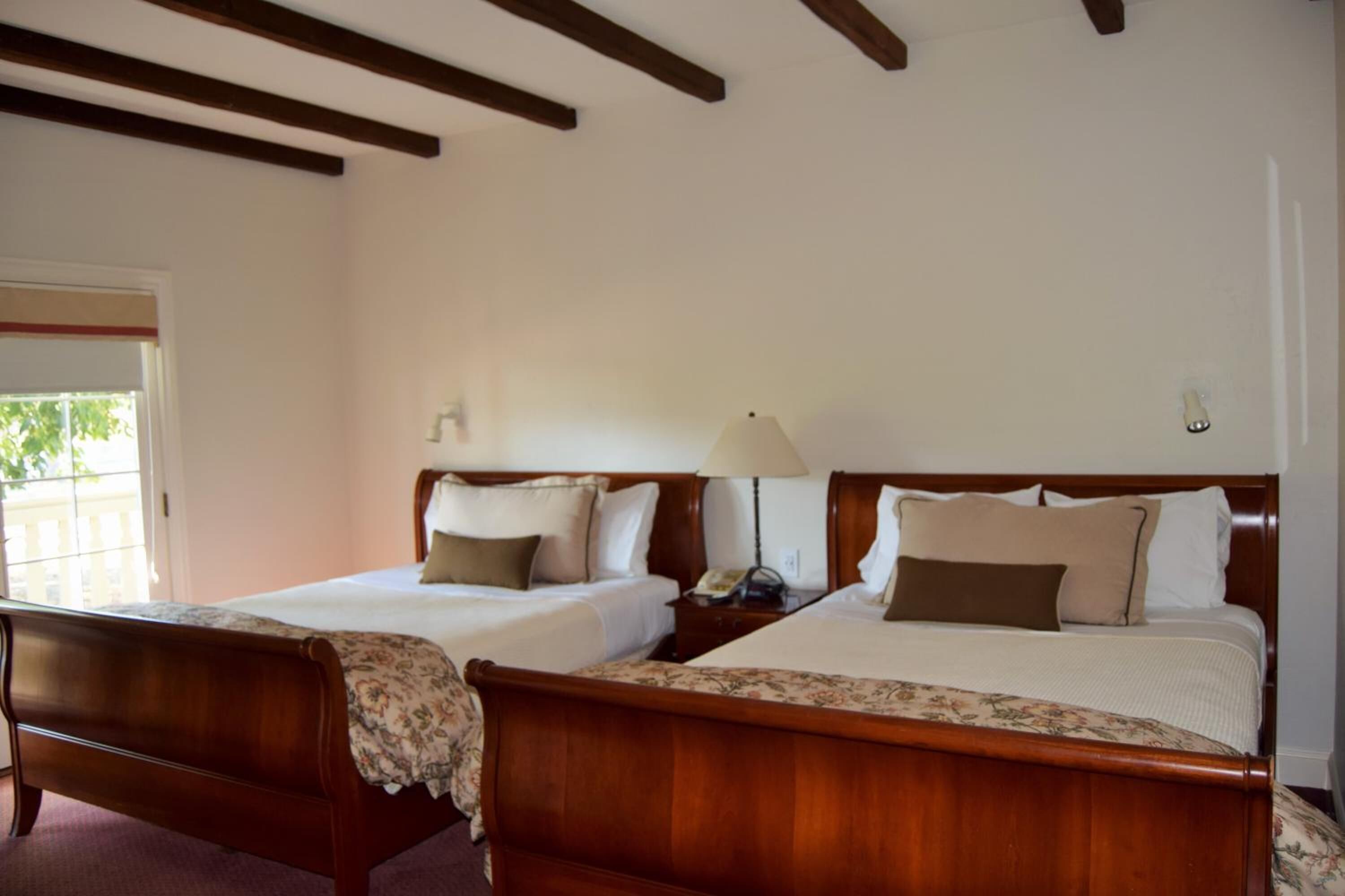 A hotel room with two wooden beds, a nightstand with a lamp, and a window with a view of greenery outside.