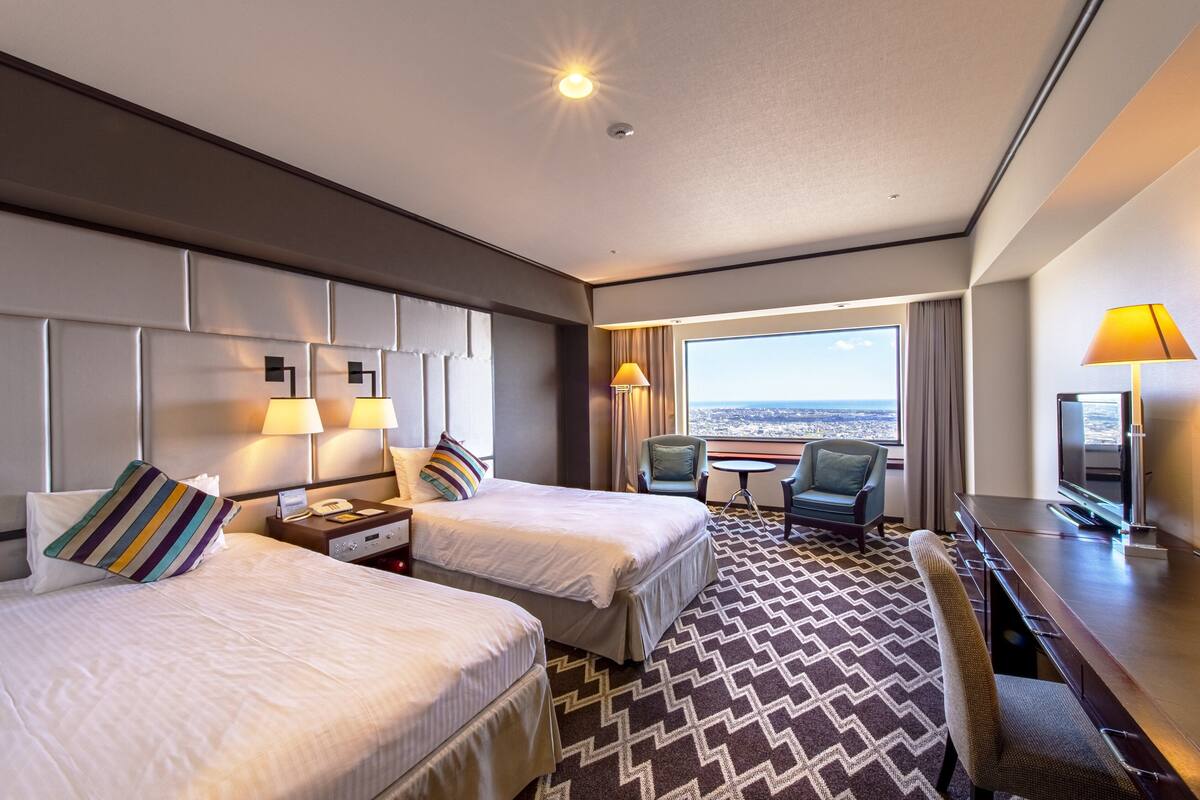 A hotel room with two beds, a desk, a chair, a TV, and a view of the city.
