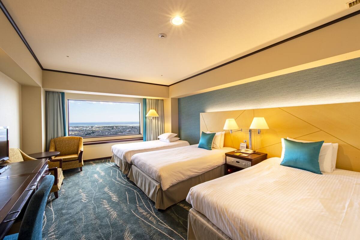 A hotel room with two beds, a desk, a chair, a TV, and a window with a view.