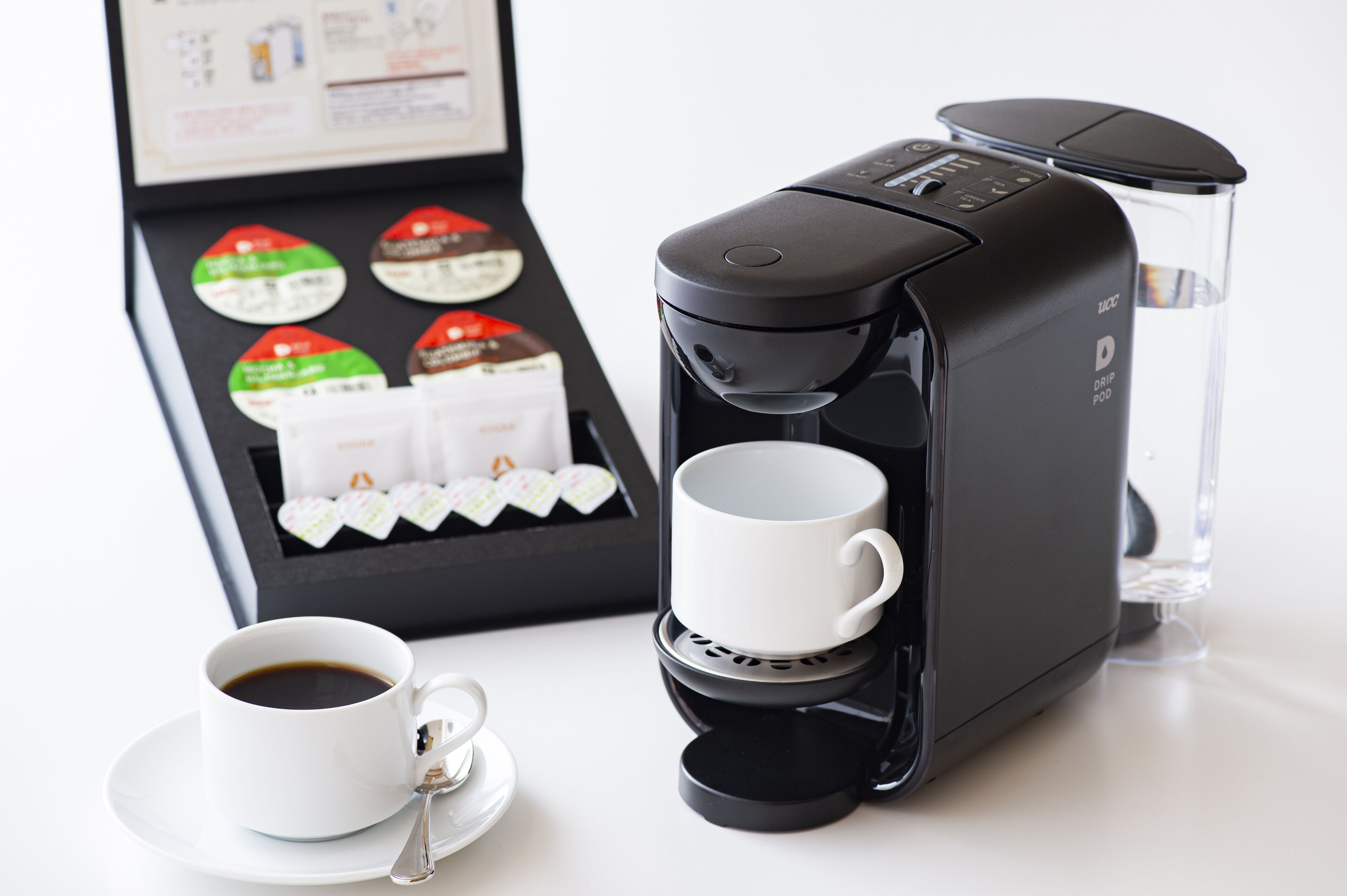 Uma cafeteira de filtro, uma chávena de café e uma caixa de cápsulas de café.