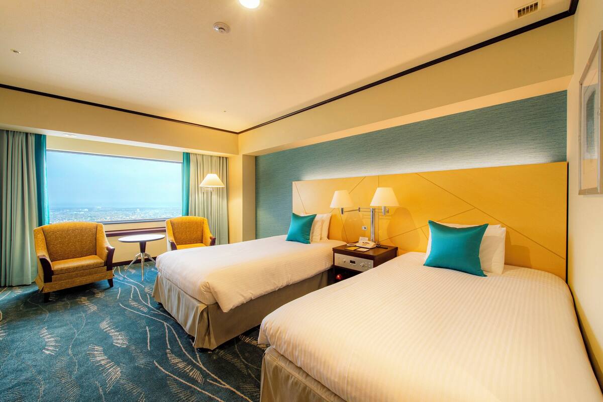 A hotel room with two beds, a chair, a small table, and a view of the city.