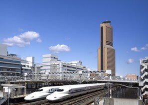 Una moderna stazione ferroviaria con due treni ad alta velocità su binari adiacenti, un ponte pedonale sopraelevato e un grattacielo sullo sfondo.