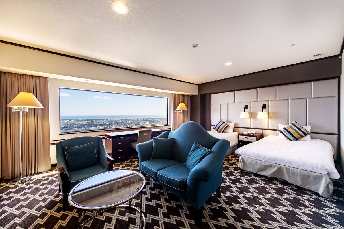 A hotel room with a large window, a sofa, a bed, and a small table.