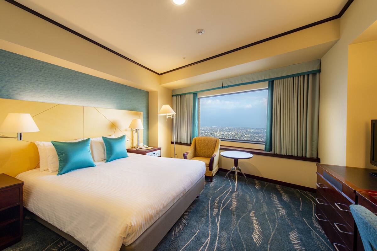 A hotel room with a large bed, two bedside lamps, a chair, a small table, a TV, and a window with curtains.