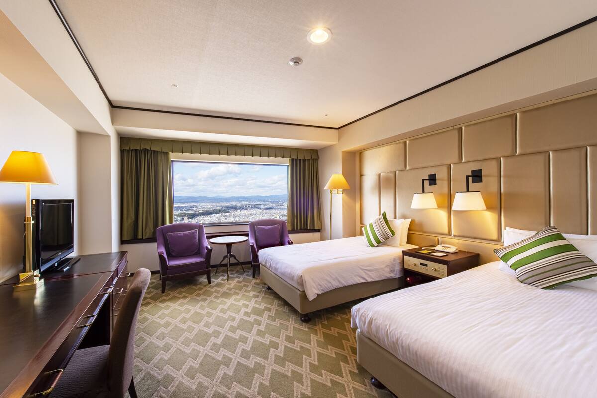 A hotel room with two beds, a desk with a TV, a small table, and a view of the city.